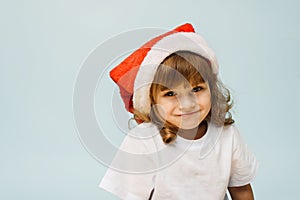 Funny cute girl in a santa hat stands on a blue background with copy space. Concept of christmas and new year.