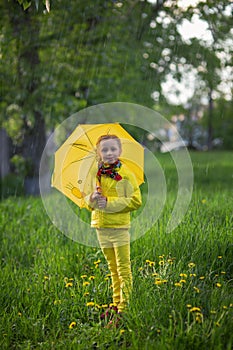 Funny cute girl wearing yellow coat holding colorful umbrella playing in the garden by rain and sun weather on a warm autumn or