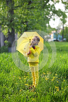 Funny cute girl wearing yellow coat holding colorful umbrella playing in the garden by rain and sun weather on a warm autumn or