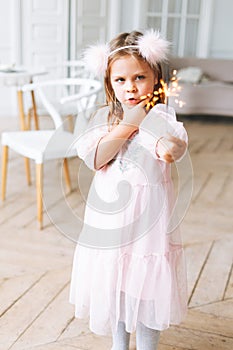 Funny cute little girl with long hair in light pink dress holding burning sparkler candle in hands in bright living room at the