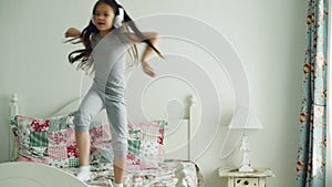 Funny cute little girl in wireless headphones dancing and have fun on bed at home in cozy bedroom
