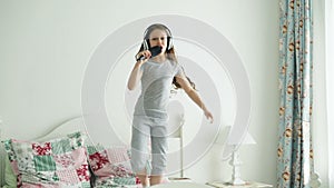 Funny cute little girl in wireless headphones dancing singing with comb and have fun on bed at home in cozy bedroom