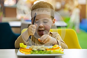 A funny cute three year old toddler boy eats fresh fruit while sitting at a table in a public place