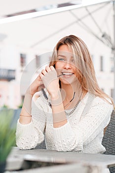 Funny cute young blond woman in a vintage knitted white sweater is sitting at a table in a street cafe. Beautiful girl on vacation