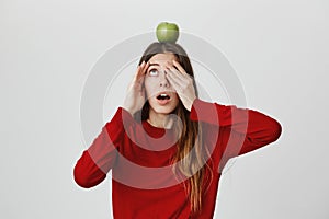 Funny cute young girl in red top covering one eye while peeking on apple she holds with head, looking shocked and