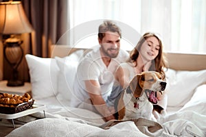 Funny dog yawns while sits in bed near hosts, satisfied with delicious breakfast. Happy female and make in background