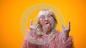 Funny elderly woman in pink coat making rocker gestures and showing tongue, fun