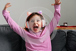 Funny emotional child girl with pink ears wireless headphones at home on the sofa.