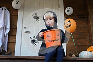 Funny emotional little child in halloween costume sitting on porch of house, holding trick or treat candy basket and surprisely