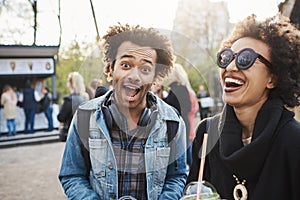 Funny emotive young dark-skinned male with afro haircut making face at camera while spending time in park with best