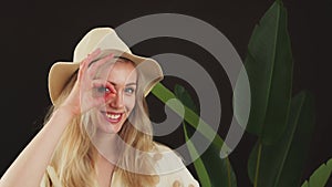 Funny European blonde young girl standing in front of a big-leaf plant in a hat doing ok gesture with hand smiling, eye