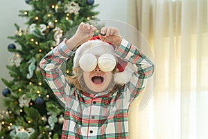 Funny excited child with Christmas balls on eyas near Christmas tree at home. Child preparing for the Christmas and New