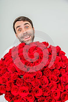 Funny expressive man isolated on white background, holding huge bunch of roses and looking from behind them with really