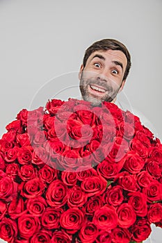 Funny expressive man  on white background, holding huge bunch of roses and looking from behind them with really