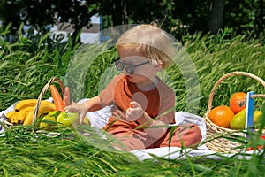 funny fair-haired boy with sunglasses holds an apple, basket with fruits on green grass