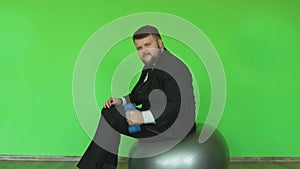 Funny fat businessman with dumbbell sits and moves on exercise ball in gym. chromakey green. Bearded thick guy in black