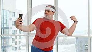 Funny fat man with big belly posing and doing selfie in bright gym or at home. Overweight male trying to look attractive