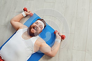 Funny fat man doing exercises with dumbbells on a workout on the floor in the house.