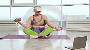 Funny fat man with dumbbells and VR glasses doing exercises in bright room. Plus size body positive with laptop at home