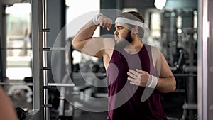 Funny fat man looking at mirror reflection gym and posing, pretending muscular