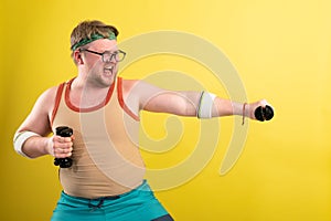 Funny fat man in sportswear with dumbbells in his hands. Overweight man goes in for sports. Yellow background isolate