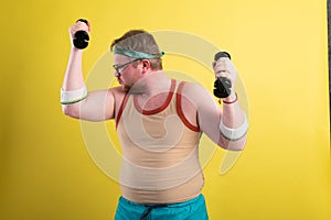 Funny fat man in sportswear with dumbbells in his hands. Overweight man goes in for sports. Yellow background isolate