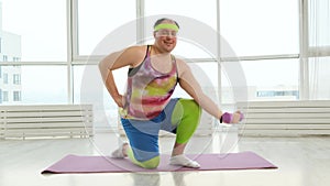 Funny fat man is training with dumbbells in bright room. Nerd overweight male in bright clothes engaged with dumbbells