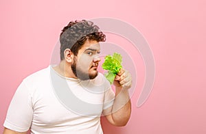 Funny fat man in a white T-shirt stands on a pink background with lettuce in his hands, sniffs fresh greens and looks at the