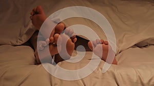 Funny feet of a kid boy and mom under a blanket on a sheet at night. Happy baby child girl and mother with bare legs