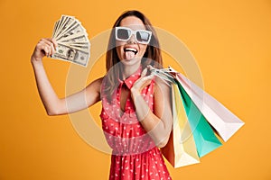 Funny female shopaholic holding fan of money and colorful shopping bags, looking at camera