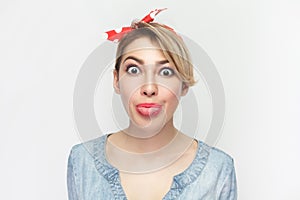 Funny foolish woman in denim shirt and red headband standing sticking tongue out, looking at camera.