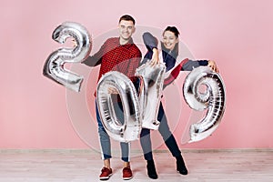 Funny friends girl and guy dressed in stylish clothes are holding balloons in the shape of numbers 2019 on a pink
