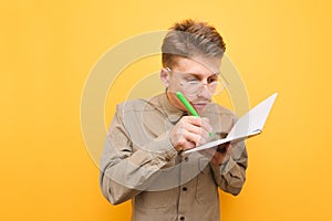Funny geek wearing glasses and a shirt stands on a yellow background and writes in a notebook in a focused manner. Funny nerd with
