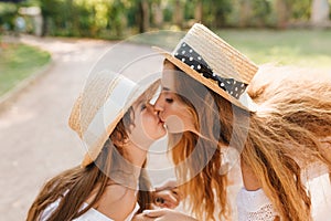 Funny girl with light-brown straight hair kissing mom during walk on the alley. Outdoor close-up portrait of lovely