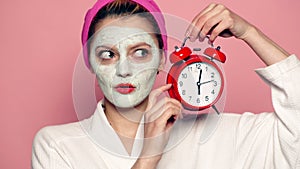 Funny girl with mask on face holds alarm clock in hand. Beautiful girl with cosmetic mask on face. Woman wearing face