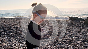 Funny girl plays with sunglasses on pebble beach at sunset