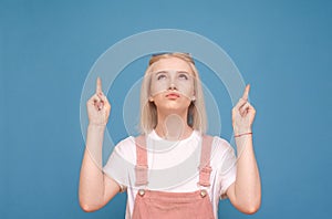 Funny girl in a pretty casual dress is standing against the background of a blue wall, looking up and showing her fingers on