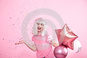 Funny Girl with silver hair gives a smile and emotion on pink background. Young woman or teen girl with balloons and confetti