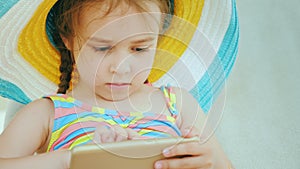 Funny girl in a striped hat playing on a smartphone, resting in a lounge chair by the pool