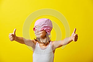 Funny girl in a swimsuit stands on a yellow background, shows thumbs up, wearing a swimming cap and glasses, portrait on