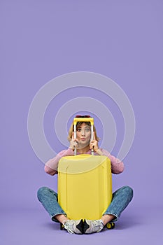 Funny girl traveler sitting with yellow travel suitcase isolated on purple.
