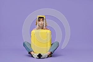 Funny girl traveler sitting with yellow travel suitcase isolated on purple.