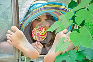 Funny girl wearing a colorful hat with lollipop and showing her feet in window with grape leaves