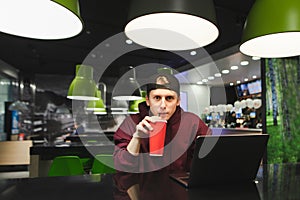 Funny guy in casual clothes sits with laptop at table in fast food cafe, drinks cool cola and looks in camera with shocked face.