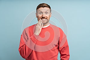 Funny guy covering mouth with hand laughs making big eyes, oops isolated on blue studio background.