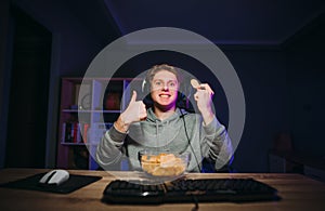 Funny guy in a headset on his head and casual clothes sits at the computer at night with a plate and chips in his hand and shows a