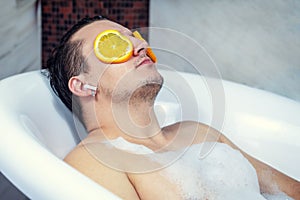 Funny guy washing his bath. Listens to music with wireless headphones. Relaxed from spa procedures on face with orange round slice
