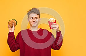 Funny guy wearing casual clothes is standing on a yellow background with French fries and a burger in his hand. The hungry young