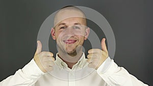 Funny handsome guy smiles and shows thumb up with both hands. Portrait on gray background.