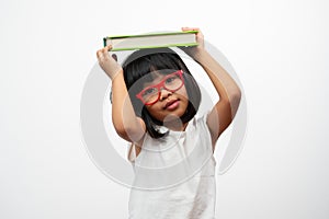 Funny and Happy Asian little preschool girl wearing red glasses holding a green book on the head, on white isolated background.